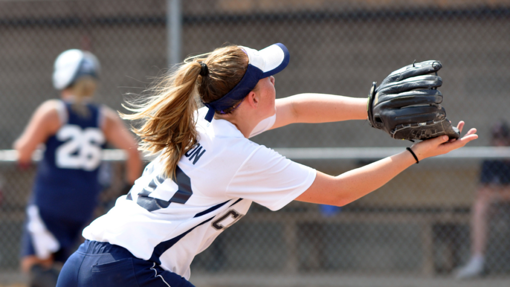 High school softball player in white and blue shirt and blue ball cap reaches out to catch a softball.