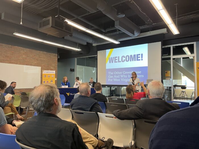 A group of people listens to a presenter at the front of the room in front of a projection screen.