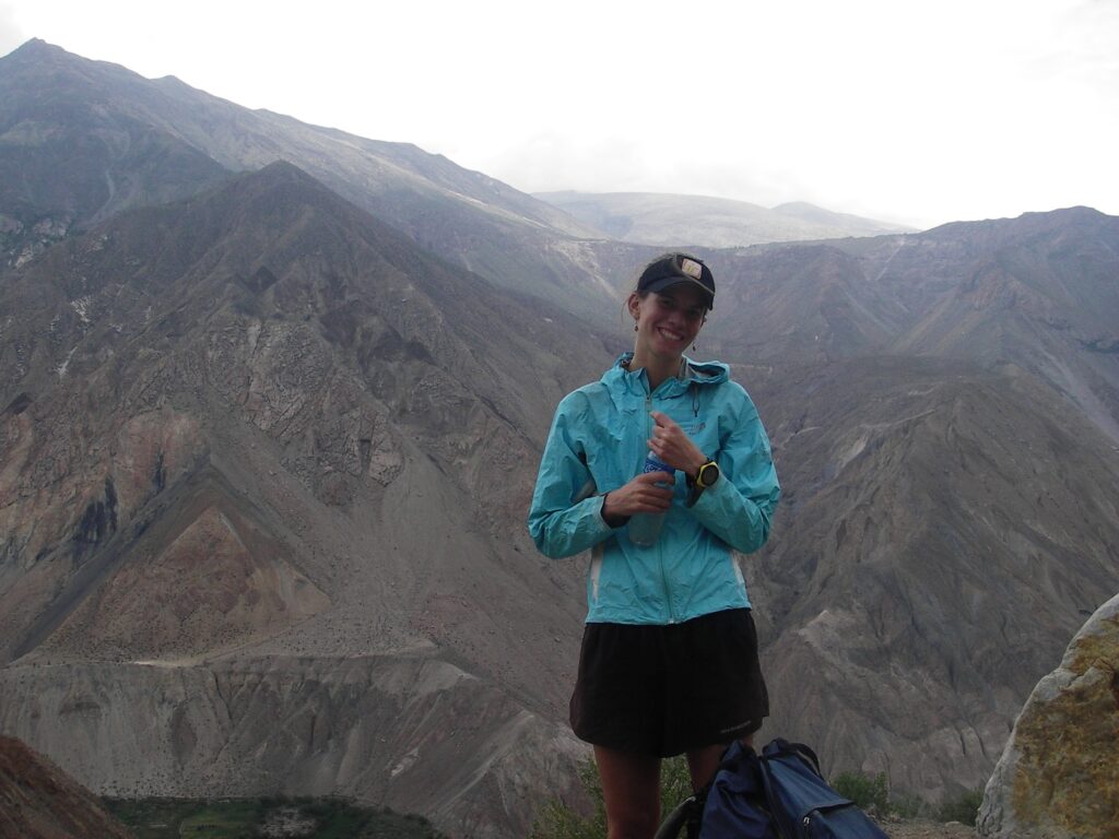 A woman smiling stands atop a mountain. She wears a blue hoodie and black shorts and wears a ball cap.