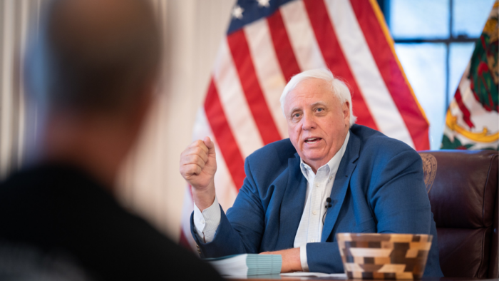 A man with white hair and wearing a blue suit jacket speaks while sitting at a table with the American flag behind him.