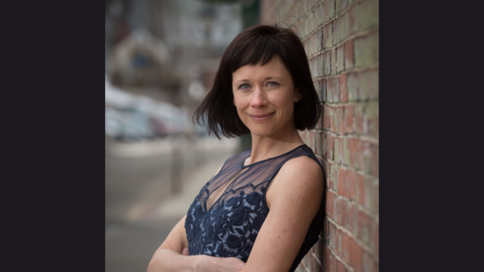 A woman in a black, sleeveless dress with short black hair stands leaning against a brick wall.