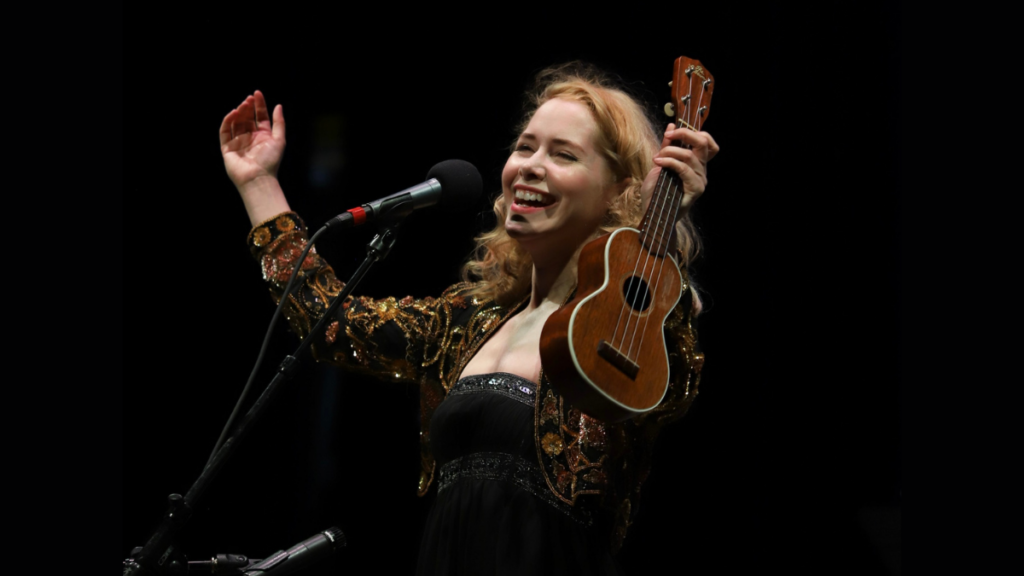 A woman stands at a microphone, arms up, smiling, and holding a ukulele.