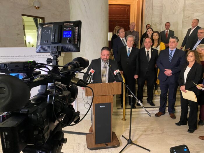 A man stands a podium and speaks into a microphone. Several other individuals stand behind him. A TV camera can be seen capturing the moment.