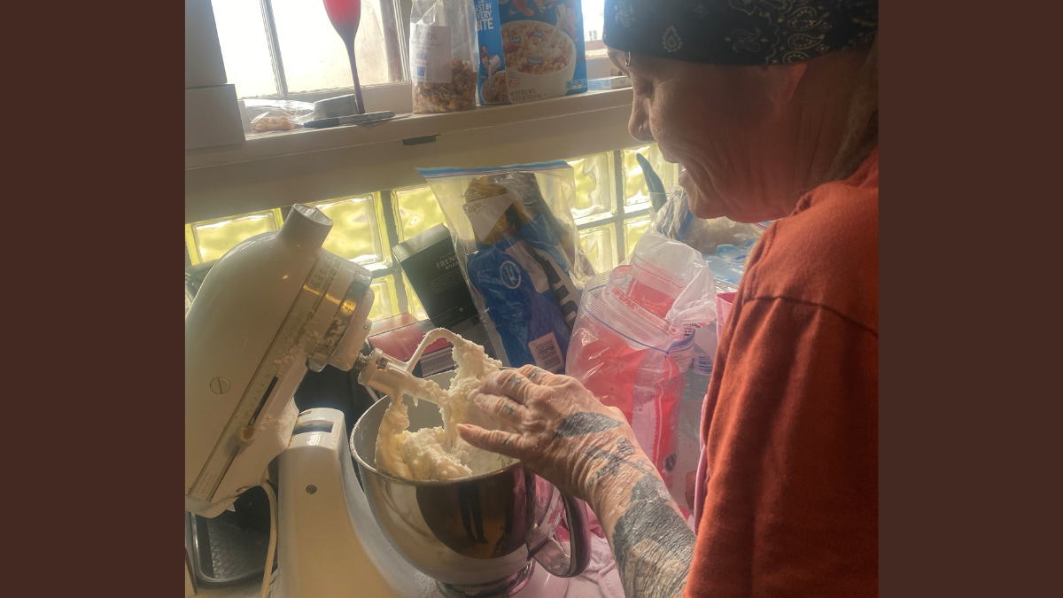 A woman in a light maroon shirt wearing a bandana on her head mashes potatoes in a mixer.