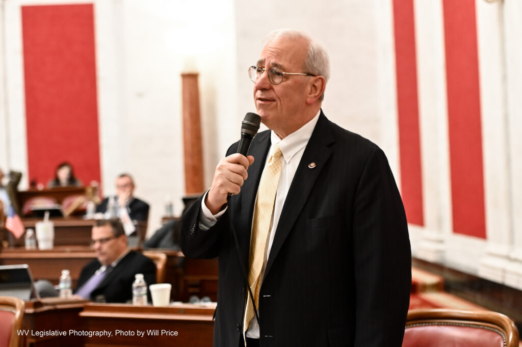 A man wearing a suit and yellow tie speaks into a microphone to a body of lawmakers.