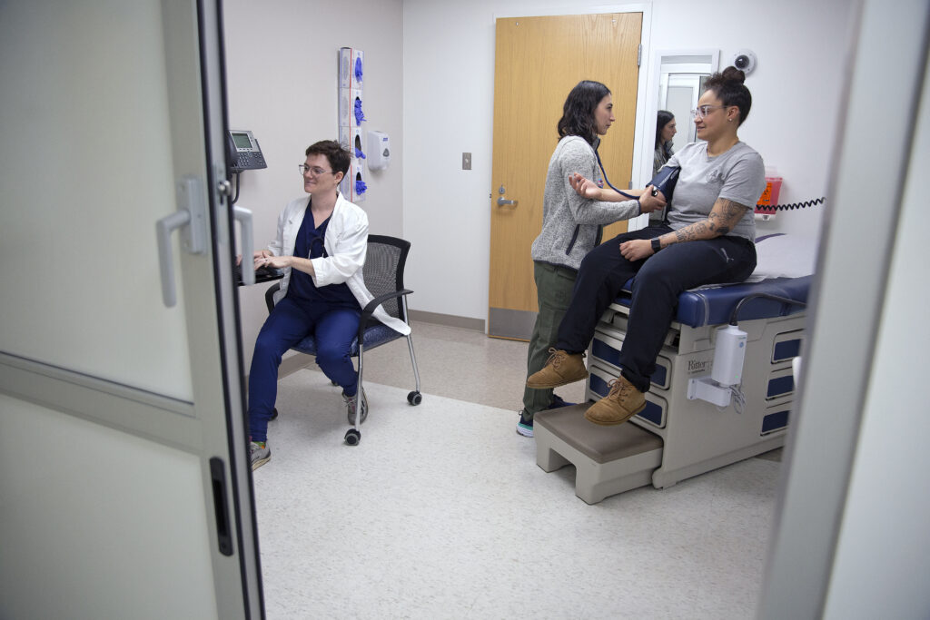 Three people can be seen in a medical room. One of the people is a patient having her vitals checked by a nurse.