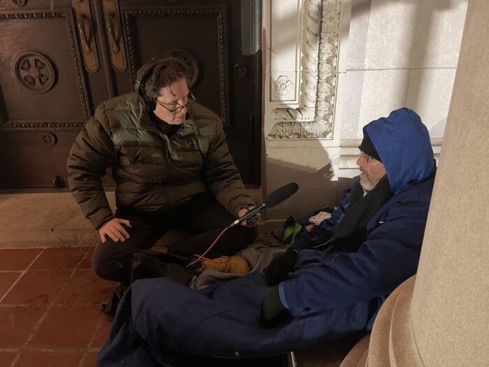 A man bends down to be on eye level with another man who is homeless. The man who has bent down is holding a microphone and wears professional headphones. The man who is bent down is conducting an interview with the homeless man.