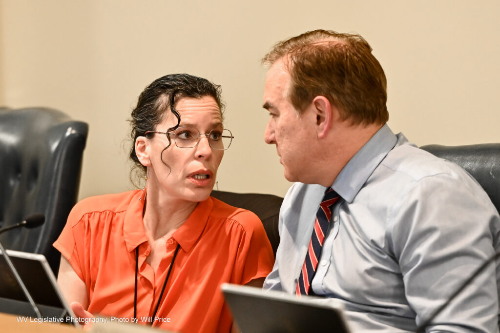 A woman in an orange shirt speaks with a man wearing a suit and tie during a Senate Judiciary meeting.