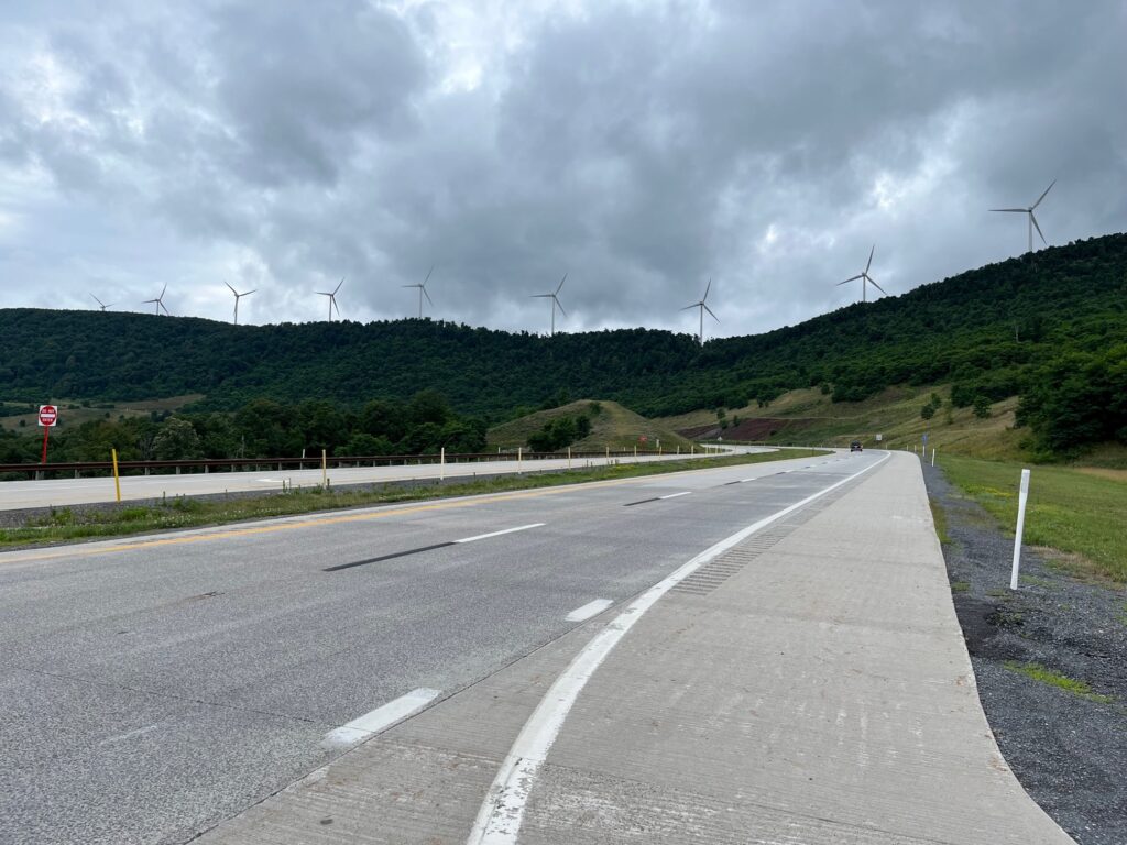 Wind turbines on a ridge tower over the deserted Corridor H highway in northern West Virginia against an overcast sky.
