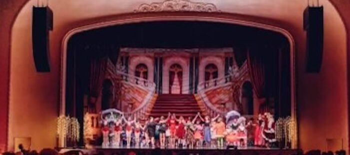 A large stage for theater is shown. A line of performers stand next to each other with their hands raised up in the air.