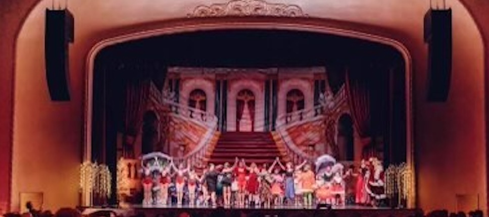 A large stage for theater is shown. A line of performers stand next to each other with their hands raised up in the air.