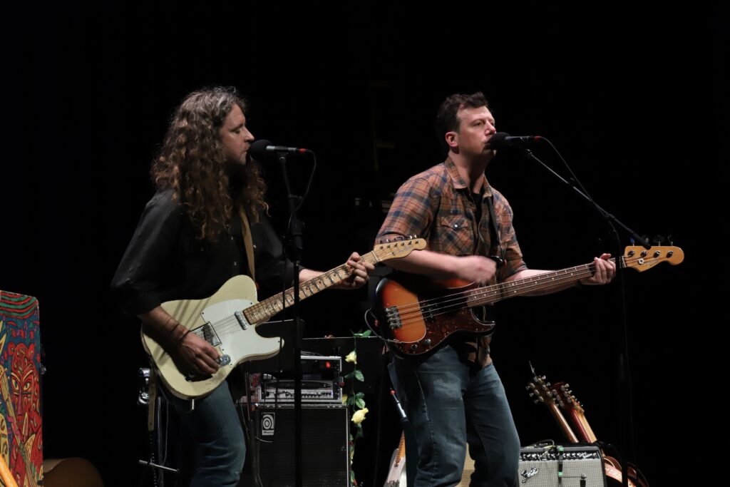 Two men stand next to each other a stage. One man plays a guitar, while the other plays a bass.