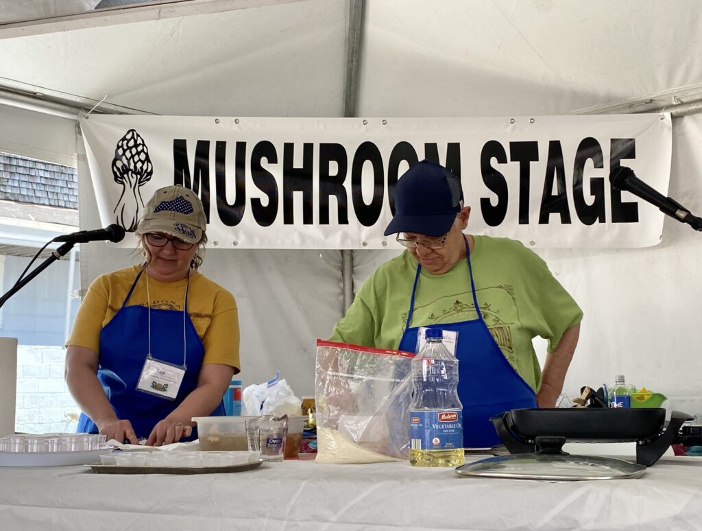 Two people stand side by side cutting mushrooms. A sign behind them reads "Mushroom Stage."