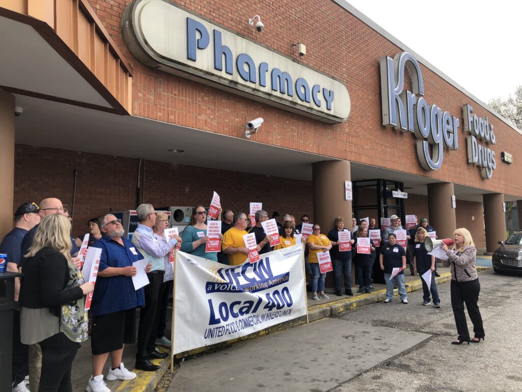 A group of union workers stand underneath a sign that says Pharmacy. The group holds a white sign with blue lettering that reads, in part, "Local 400 United Food & Commercial Workers Union" Most of the group wear blue, and some hold white signs with red lettering. One woman stands in front of the group, just below a large blue Kroger sign, wearing pink and black and holding a white and red bullhorn.