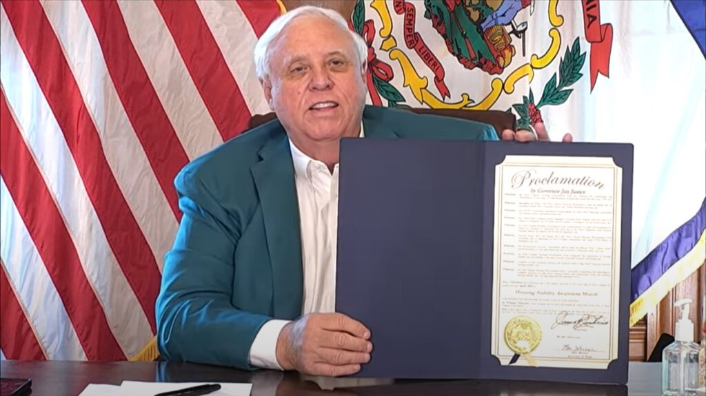 Gov. Jim Justice wears a green blazer over a white shirt while holding a blue folder that contains a proclamation. He is seated in front of the red and white stripes of the American flag, as well as the West Virginia state seal on the state flag.