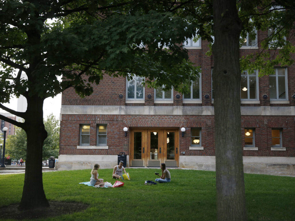 Students are seen sitting in the grass on a college campus.