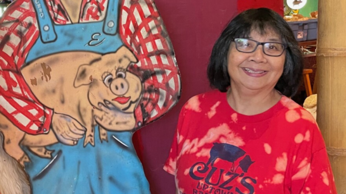 A woman in a red shirt and glasses, short black hair stands smiling next to a cut out of Elvis Presley holding a pig.