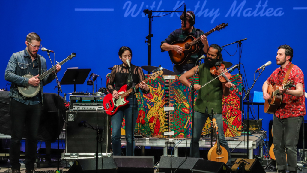 Five musicians stand on a stage playing the banjo, electric guitar, fiddle, acoustic guitar, and bass guitar.