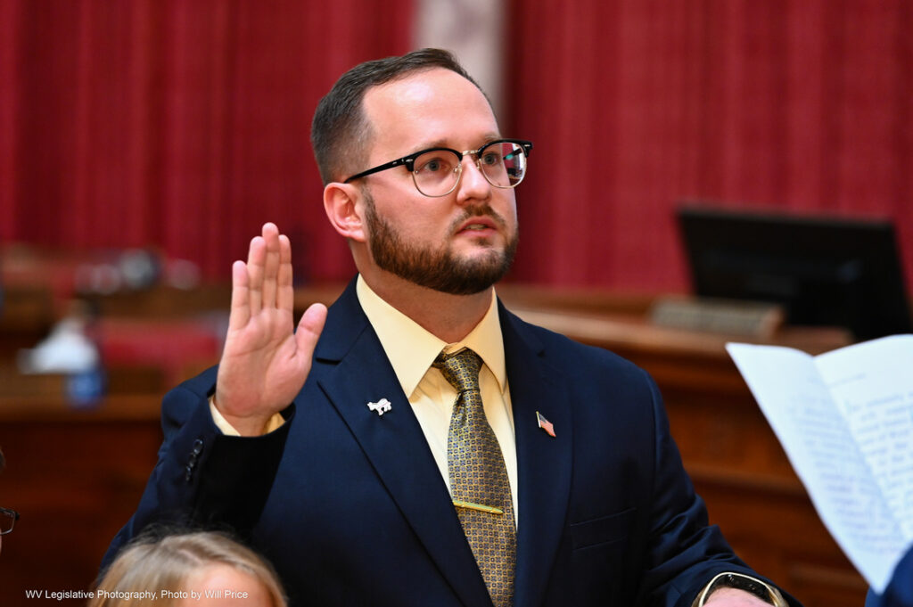 A man wearing glasses and a suit and tie with a donkey pin and American flag pin on his lapel. He raises his right hand as he takes the oath of office.