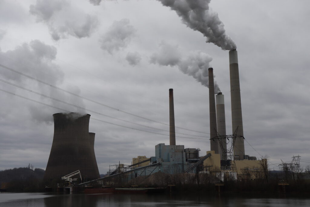 Steam and emissions rise from the tall stacks and cooling towers of the John Amos power plant against overcast skies next to the muddy water of the Kanawha River.