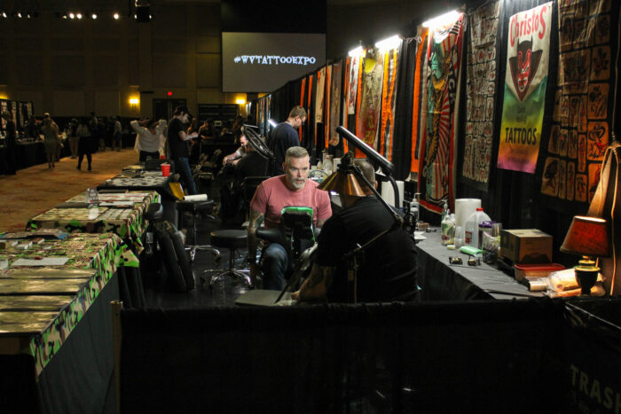 Two men speak next to vibrantly colored banners of traditional tattoo art. Behind them can be seen more booths, and in the far background a sign reading #WVTattooExpo can be seen.