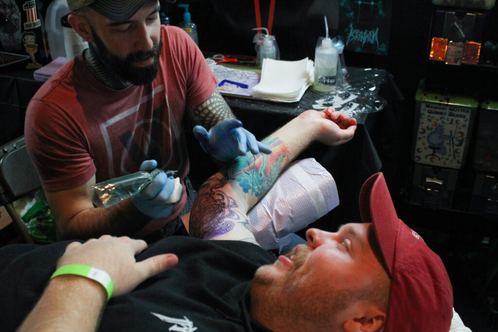 A bearded man wearing a red shirt, ball cap and black gloves holds a tattoo machine over another man's bicep. The second man, also bearded and wearing a red cap and black shirt, has a jellyfish design painted on his arm as a template for the tattoo he is receiving.