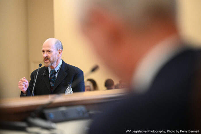 A man wearing a black suit speaks to a room of lawmakers from a podium.