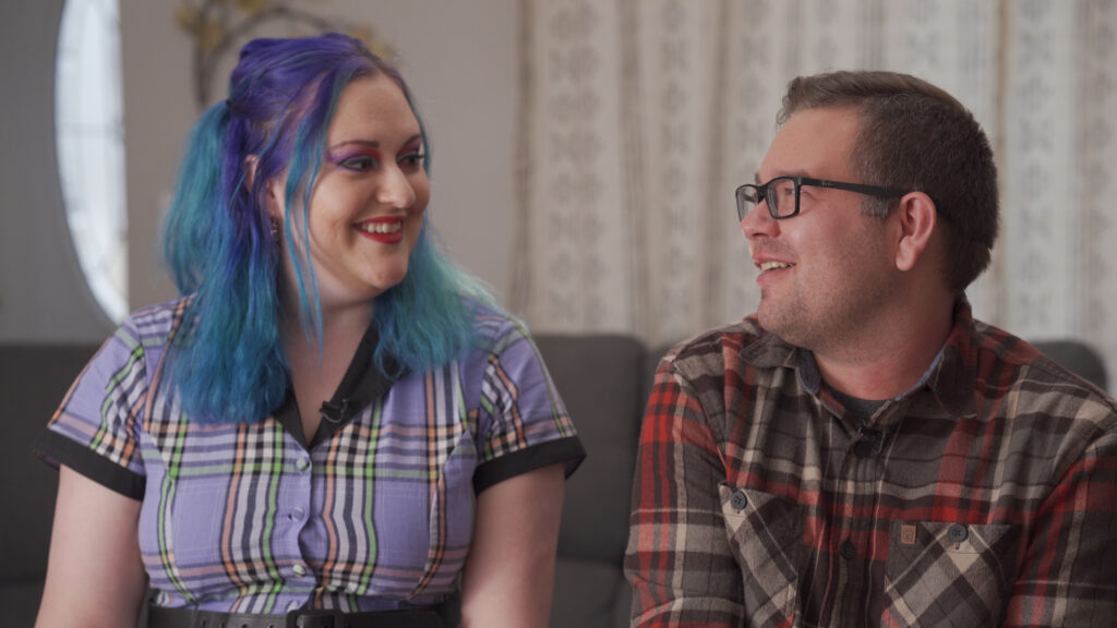 A woman with purple hair talks to a man with glasses.