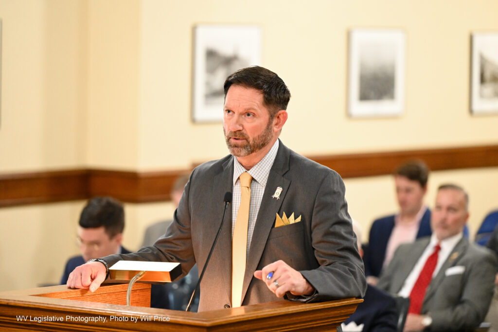 A man dressed in a suit and tie speaks from a podium in a committee room of the West Virginia Legislature.