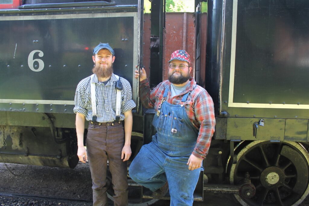Two young man stand next to each other in front of a train. One is dressed in blue overalls, while the other is dressed in brown pants with suspenders. Both are wearing ball caps.