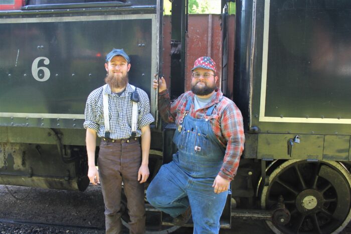Two young man stand next to each other in front of a train. One is dressed in blue overalls, while the other is dressed in brown pants with suspenders. Both are wearing ball caps.