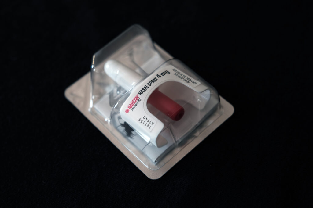 A small nasal spray is shown in medical packaging. The device is while except a red switch on the bottom.