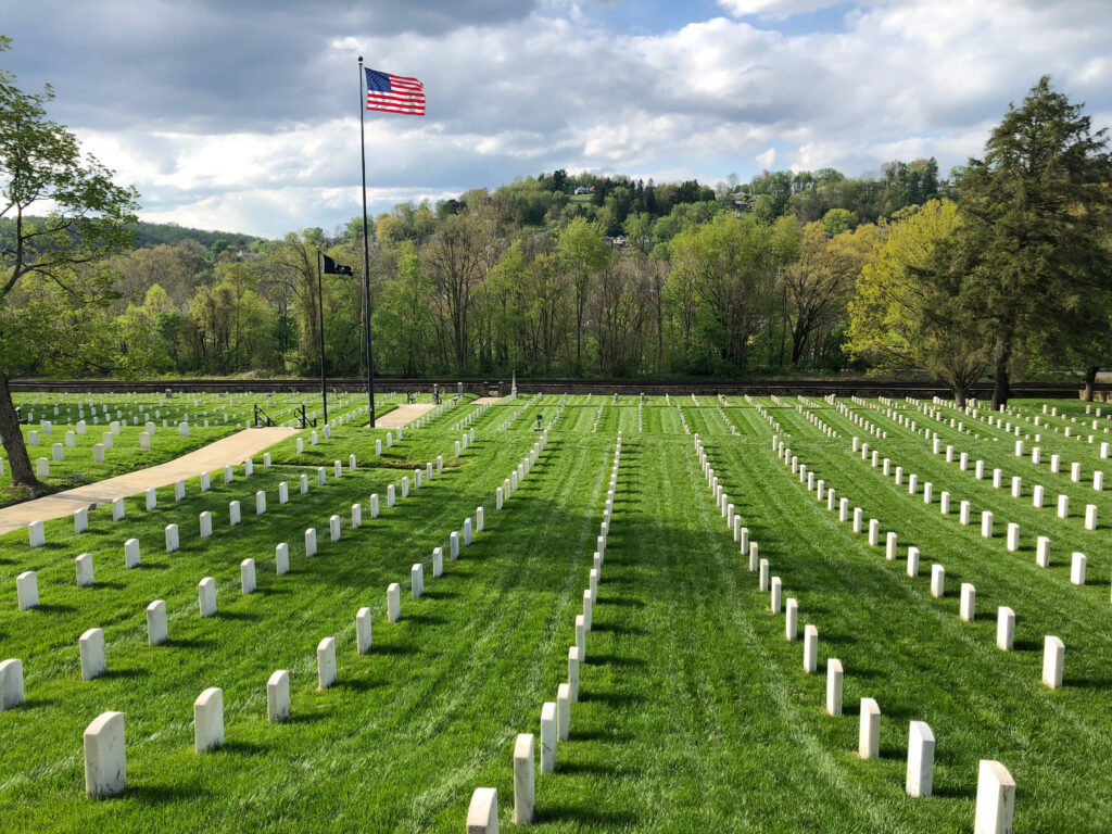 An American flag flies high over headstones at the Grafton National Cemetery. Green lanes can be seen between the grey-white stones, neatly organized in rows stretching away from the viewer. The shadow of a tree that is just visible on the left of frame is cast long over the center of frame. Trees rise on a slope in the background, beyond the cemetery below an overcast sky of grey-white clouds.