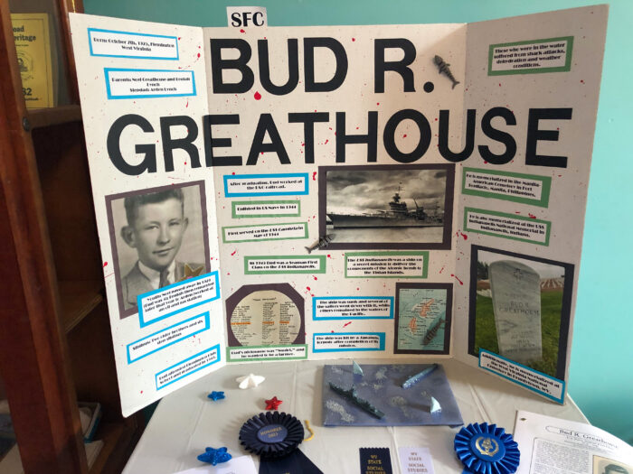 A trifold white posterboard reads "Bud R. Greathouse" and presents several facts about the man's life. To the right is a photograph of Bud, and center below the title is a photograph of the USS Indianapolis. Bottom right is a photograph of Bud's headstone in the West Virginia National Cemetery.