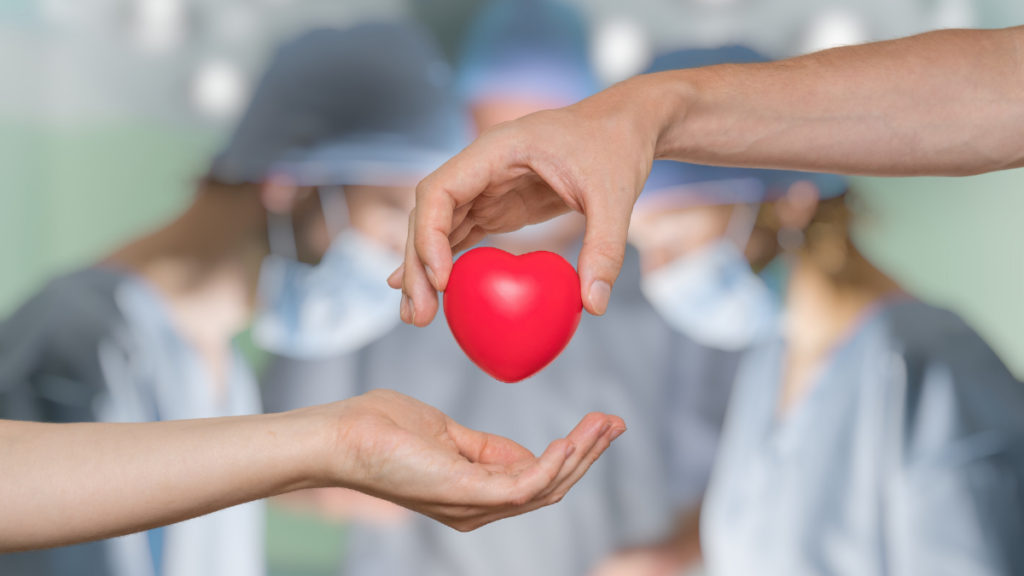 Two hands reach for each other. One hand offers a cartoon heart-shaped ball. Two doctors appear out of focus in the background and are dressed as if operating on a patient.