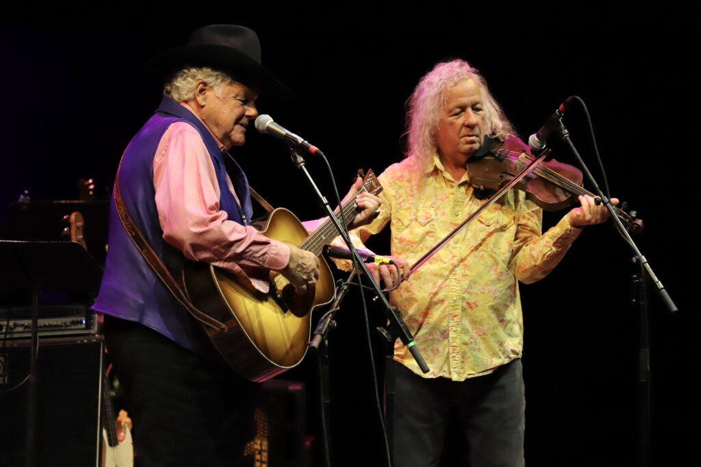 Two elderly men play instruments on stage. One man plays a guitar, and the other plays a fiddle.
