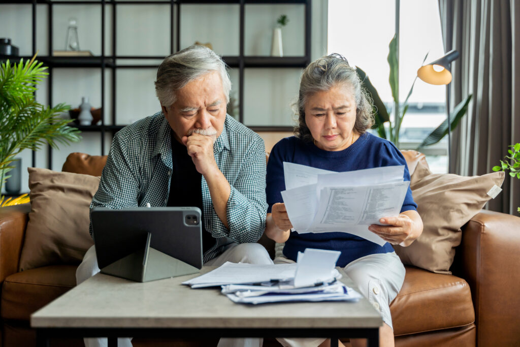 Older Asian couple sits on a couch looking over financial documuments.