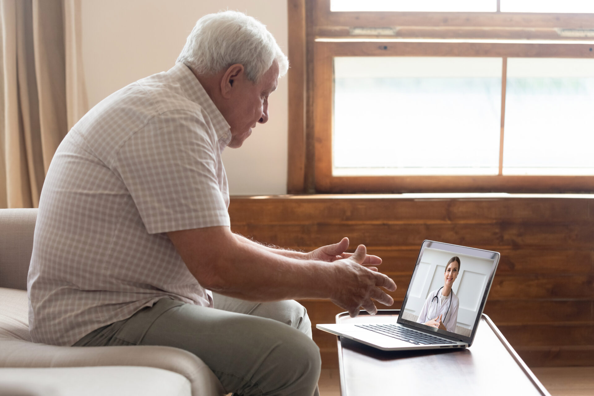 An elderly man wearing a white, checkered shirt attends a virtual doctor's appointment in a video call with a laptop.
