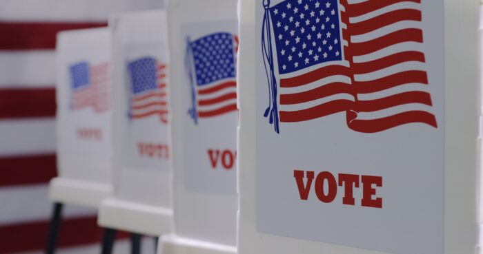 A row of voting booths. An image of the American flag can be seen on each booth with large printed letters beneath each flag that reads "VOTE" in all caps.