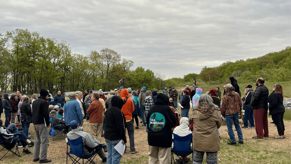 More than a dozen protesters can be seen next to trees and against a cloudy sky.