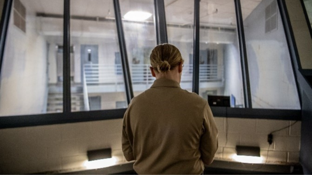 A female guard sits in a jail cage, high above inmate cells.