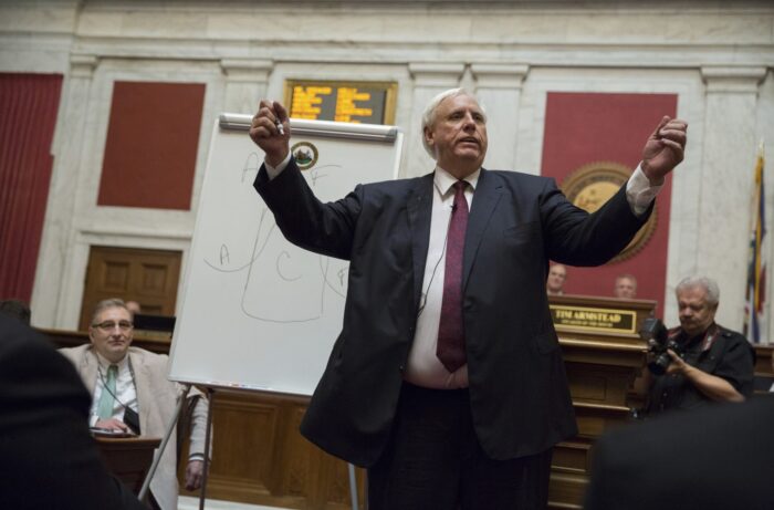 Large man raises arms, speaking to a crowd and dressed in formal attire.