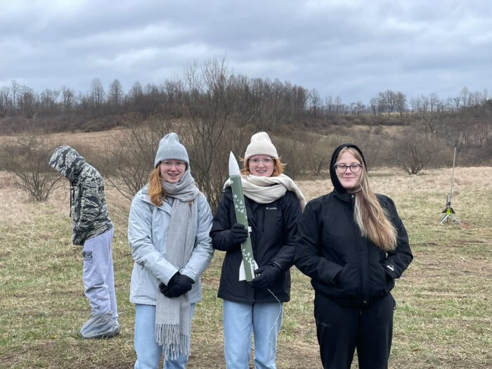 Three young women stand facing the camera wearing coats and scarves. Another person is in the background. There are trees without leaves in the distance against a cloudy sky.