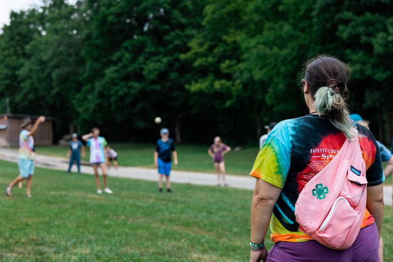 An adult wearing a tie-dye shirt and a pink bag with a green 4-H patch on it looks on as a group of children play on a green grassy field in the background.
