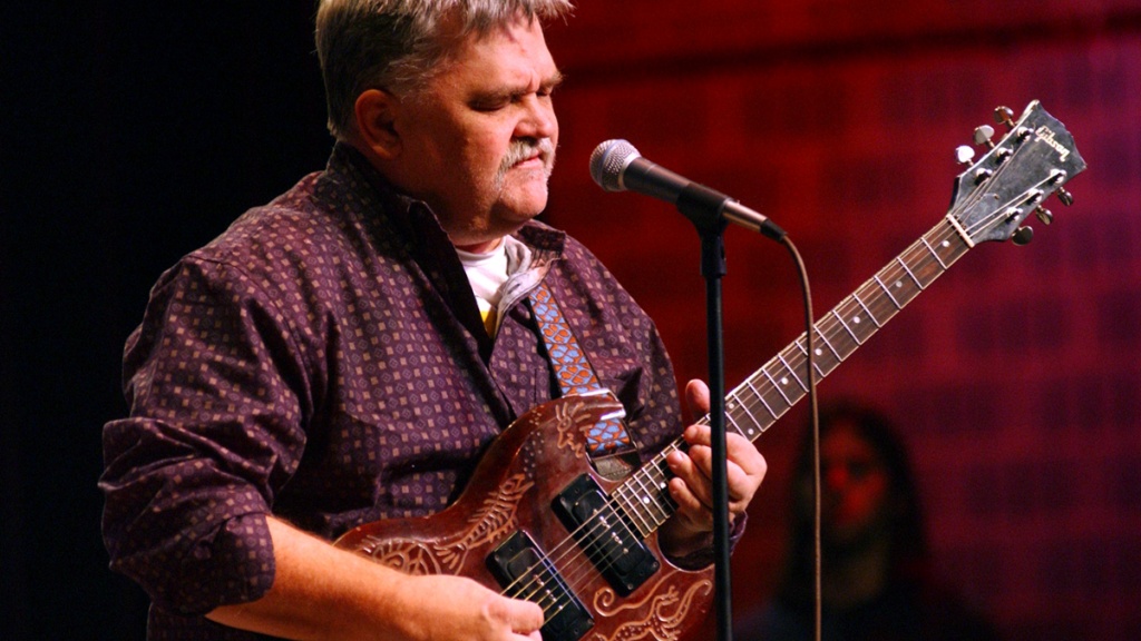 A man with grey hair and mustache plays the guitar with his eyes closed.