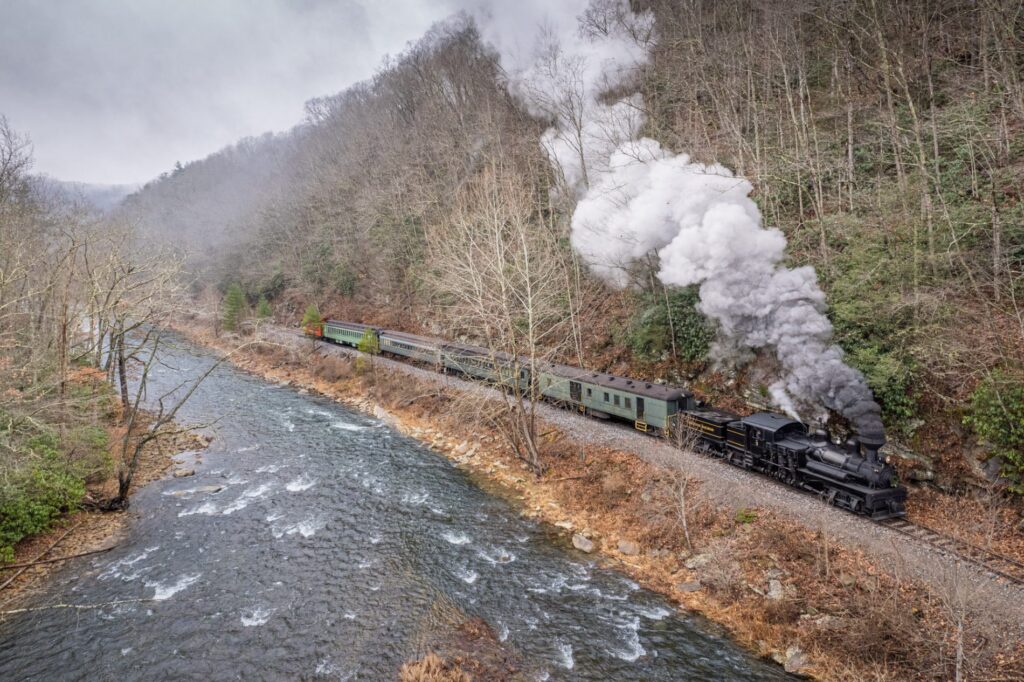 A geared steam locomotive clatters along the track, with puffs of steam streaming out of its stack against a gray sky and bare trees next to a rushing river.
