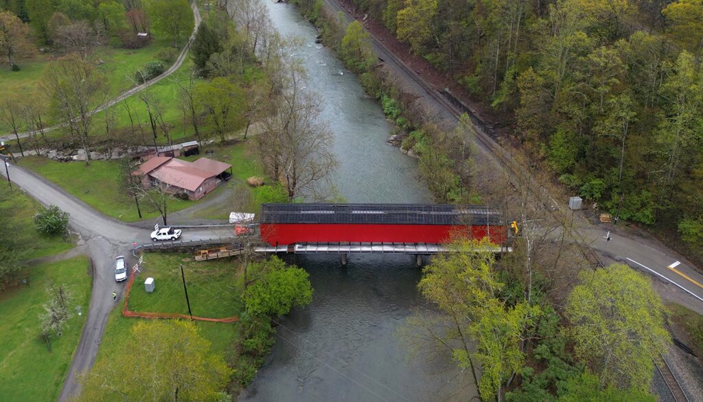 An arial image shows a covered bridge with red siding and a steel roof spanning a stream of water. To the side, heavy machinery and scaffolding can be seen.