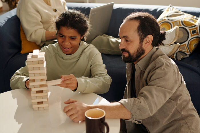 Teenage girl playing jenga with her foster dad in the living room with mother working on laptop in background