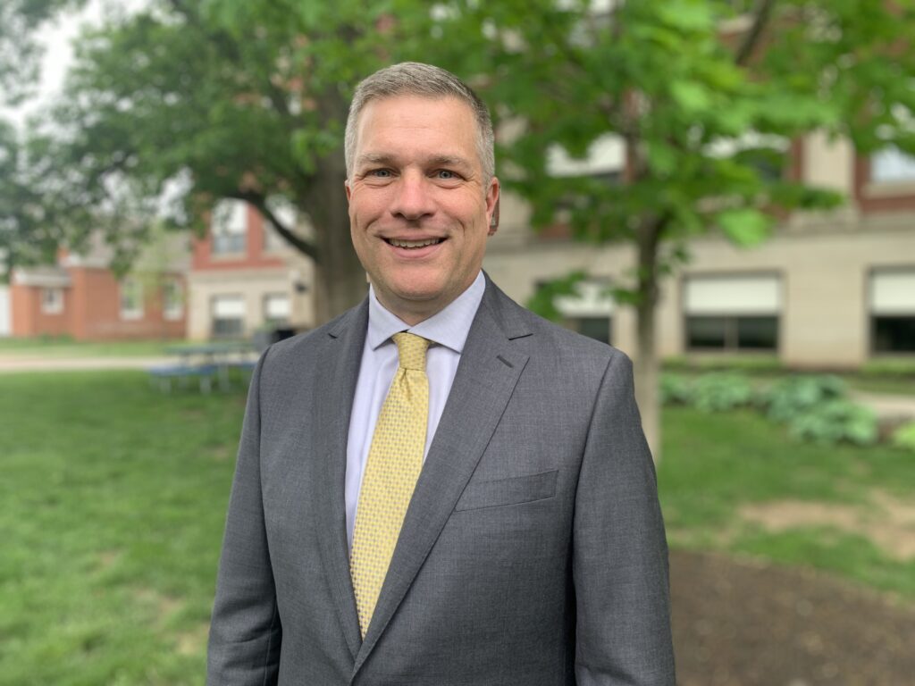 Tim Borchers wears a gray suit with a white dress shirt and yellow tie. Behind him a walkway stretches into the background flanked by a verdant lawn