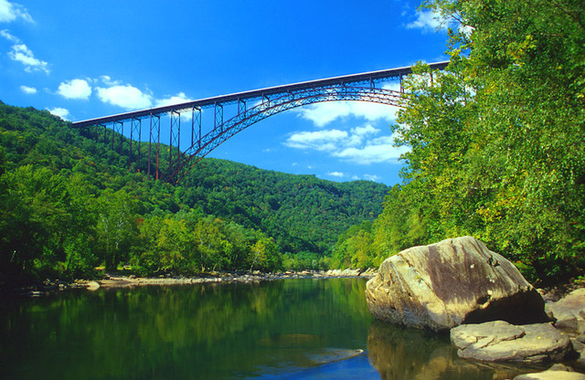 The famous arched bridge across the New River Gorge is contrasted against a bright blue sky with a few fluffy white clouds.
